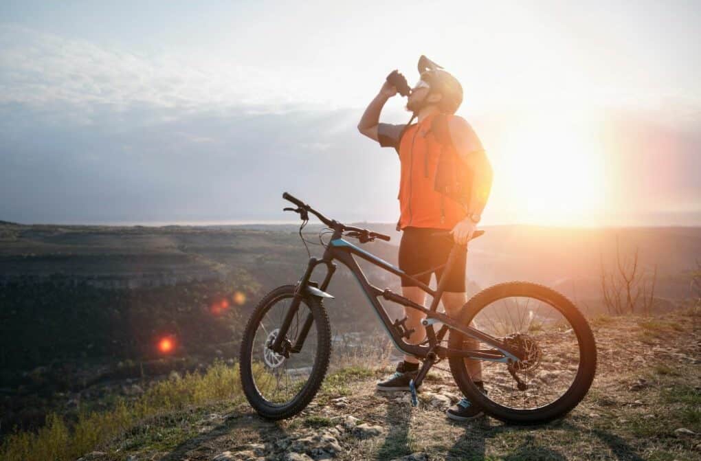 Mountain biker drinking water on the top of a mountain with a sunset behind him